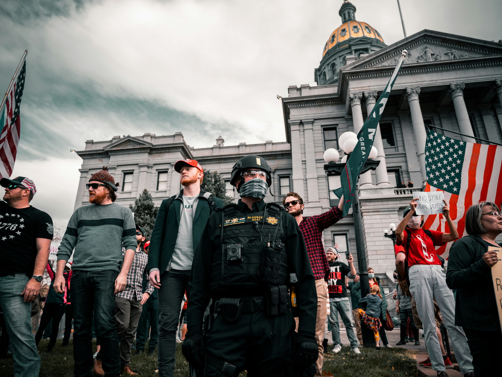 Menschenmenge mit Polizei vor Regierungsgebäude, amerikanische Flaggen und Protestschilder sichtbar