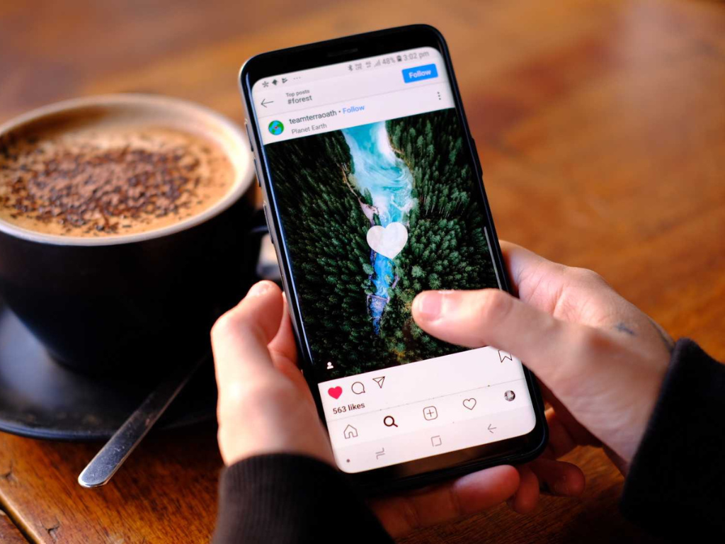 Person scrollt durch Instagram-Feed mit Naturfoto auf Smartphone – Symbolbild für Social Media Einfluss und Like-Kultur bei Kaffee im Café.