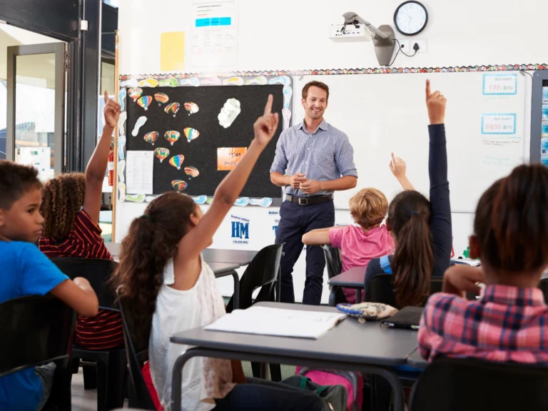 Lehrer steht vor Schulklasse, mehrere Kinder melden sich im Unterricht