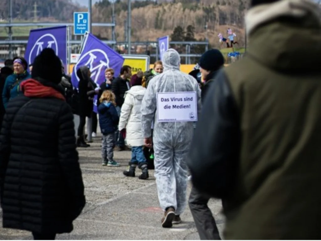 Demonstration mit Person im Schutzanzug, trägt Schild „Das Virus sind die Medien!“