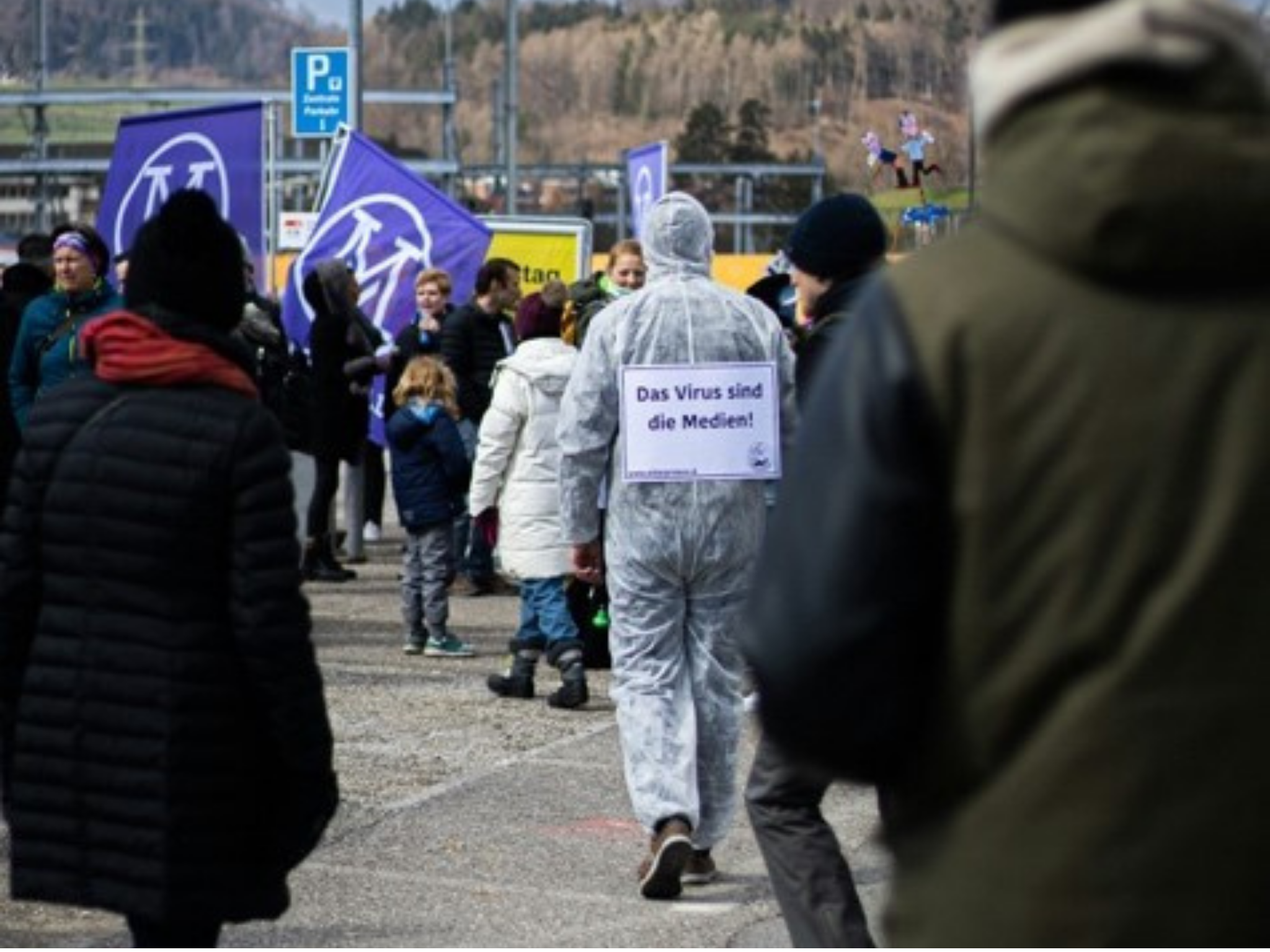 Demonstration mit Person im Schutzanzug, trägt Schild „Das Virus sind die Medien!“