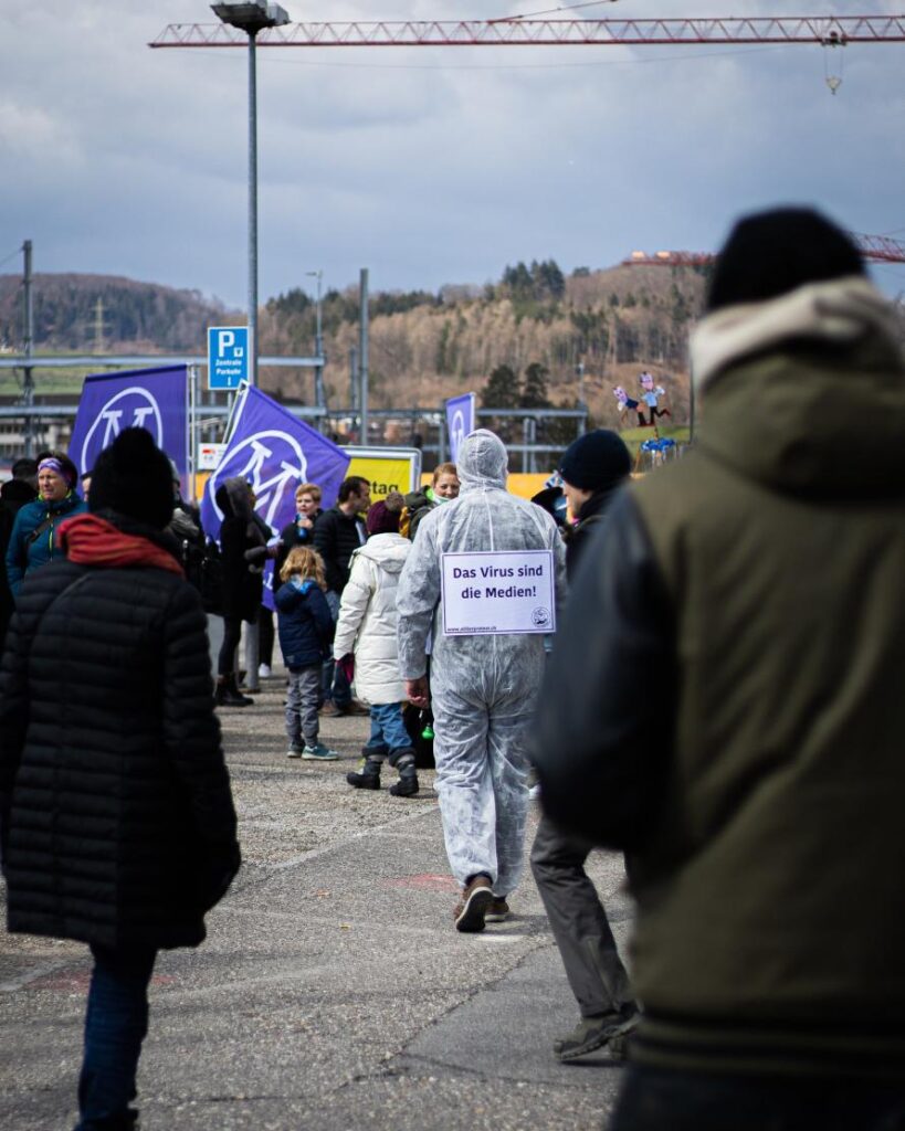 kajetan-sumila-uh05oqrt5lq-unsplash-890x-q75 Demonstration mit Person im Schutzanzug, trägt Schild „Das Virus sind die Medien!“